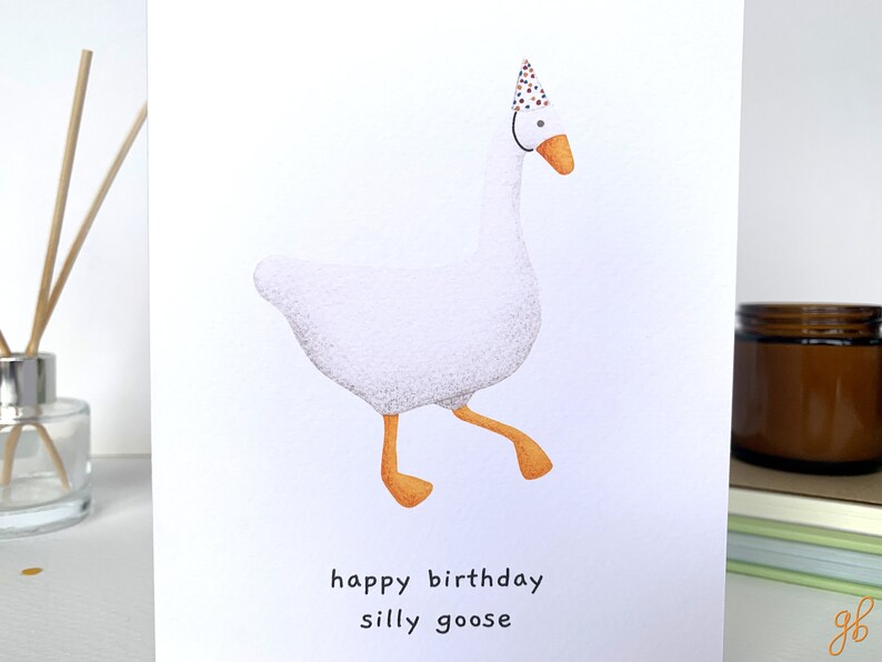 K&ouml;nnte beinhalten: Eine wei&szlig;e Gans mit einem bunten Partyhut und dem Text "happy birthday silly goose" auf wei&szlig;em Hintergrund.