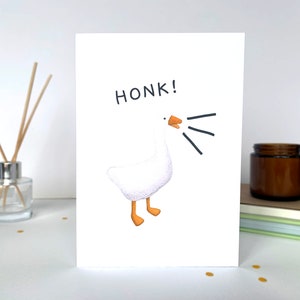 Może przedstawiać: Biała kartka z ilustracją z kresk&oacute;wki przedstawiającą gęś i słowem "HONK!" w kolorze czarnym. Gęś ma pomarańczowe stopy i dzi&oacute;b, z czarnymi liniami wskazującymi dźwięk. Kartka leży na białej powierzchni.