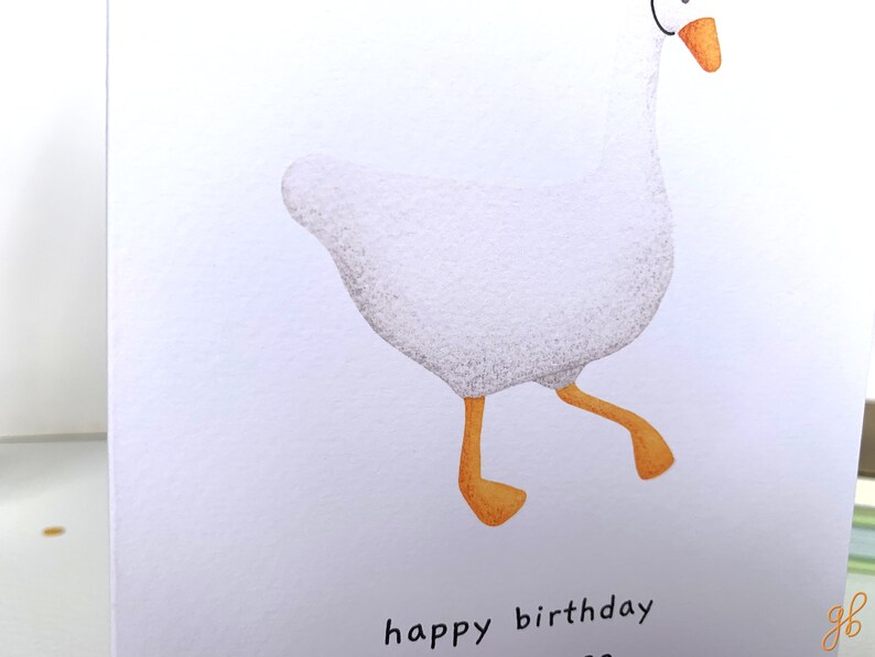 K&ouml;nnte beinhalten: Eine wei&szlig;e Gans mit orangefarbenen Beinen und Schnabel geht mit einer Happy Birthday-Botschaft auf wei&szlig;em Hintergrund.