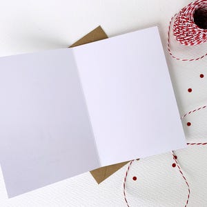 May include: An open, blank greeting card with a brown envelope underneath. A spool of red and white twine and red confetti dots are arranged on a white textured surface, suggesting a craft or stationery theme.