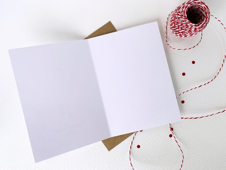 May include: An open, blank greeting card with a brown envelope underneath. A spool of red and white twine and red confetti dots are arranged on a white textured surface, suggesting a craft or stationery theme.