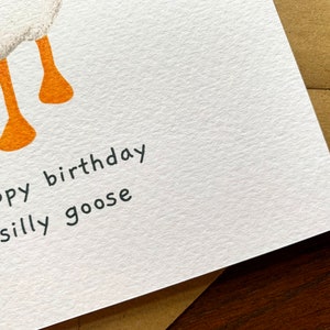 May include: A white card with the text "happy birthday silly goose" written in black ink. The card has a white goose illustration with orange legs.