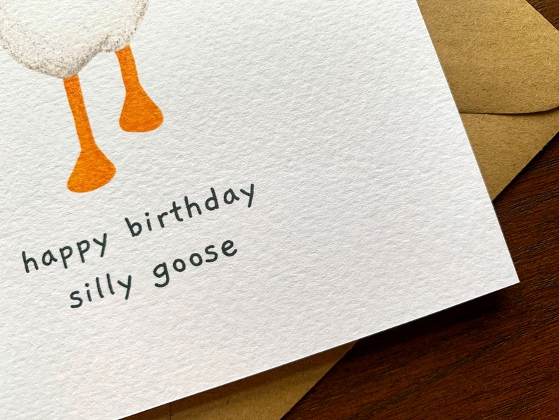 May include: A white card with the text "happy birthday silly goose" written in black ink. The card has a white goose illustration with orange legs.