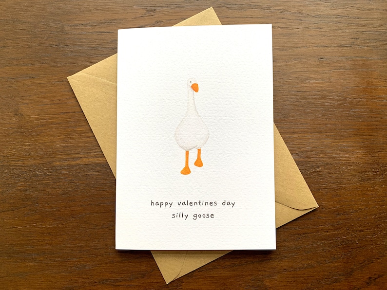 May include: A white card featuring a cartoon goose illustration and the text "happy valentines day silly goose." The card is placed inside a brown envelope, set against a wooden background.
