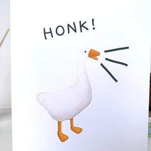 Może przedstawiać: Biała kartka z pozdrowieniami z akwarelową ilustracją gęsi z otwartym dziobem i napisem "HONK!" w kolorze czarnym. Gęś ma pomarańczowe nogi i dzi&oacute;b, z czarnymi liniami wychodzącymi z pyska.