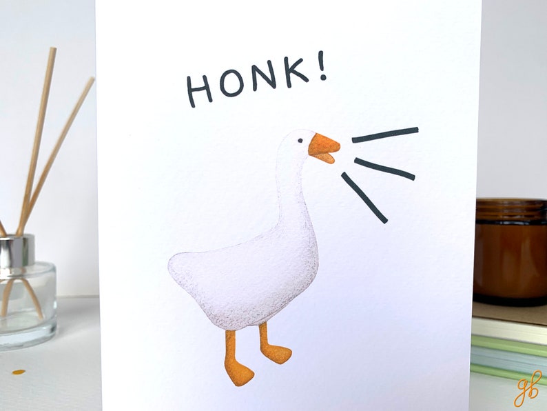 Może przedstawiać: Biała kartka z pozdrowieniami z akwarelową ilustracją gęsi z otwartym dziobem i napisem "HONK!" w kolorze czarnym. Gęś ma pomarańczowe nogi i dzi&oacute;b, z czarnymi liniami wychodzącymi z pyska.