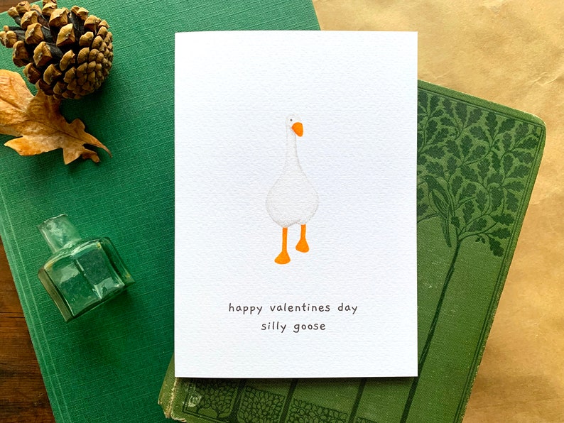 May include: A white card featuring a cartoon goose with orange feet and beak. The text "happy valentines day silly goose" is printed below the illustration. The card rests on a green book, with other decorative items nearby.