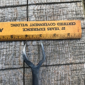 Może przedstawiać: Czarne metalowe narzędzie z rozwidlonym końcem, o długości około 12,7 cm. Narzędzie leży na drewnianej powierzchni z linijką w tle. Linijka jest oznaczona tekstem "27 Years Experience Certified Government Welding".