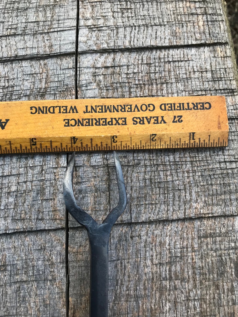 Może przedstawiać: Czarne metalowe narzędzie z rozwidlonym końcem, o długości około 12,7 cm. Narzędzie leży na drewnianej powierzchni z linijką w tle. Linijka jest oznaczona tekstem "27 Years Experience Certified Government Welding".