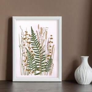 May include: A white frame with a pressed flower arrangement featuring a fern, grasses, and small brown flowers on a light pink background.