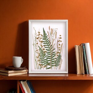 May include: A white framed print of a pressed fern and other dried plants on a white background. The print is on a wooden shelf with books on either side.