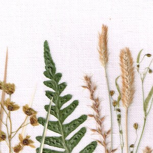 May include: A close-up of pressed botanicals on a white canvas background. The botanicals include a fern frond, a tall grass, and a smaller grass with seed heads.