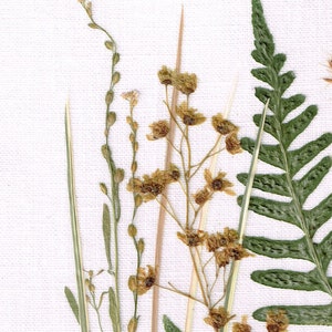 May include: A pressed flower arrangement featuring a variety of dried plants, including ferns, grasses, and delicate flowers with brown petals. The arrangement is displayed on a white linen background.