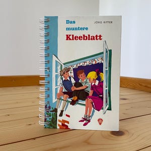 Könnte beinhalten: Ein spiralgebundenes Notizbuch mit blauem Einband und dem Titel "Das muntere Kleeblatt" in Rot und Blau. Das Cover zeigt eine Illustration von drei Kindern, die in einem Fenster sitzen. Das Notizbuch liegt auf einer Holzoberfläche.
