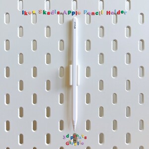 May include: A white Apple Pencil is held in place by a white plastic holder on a white pegboard with the text "IKEA Skadis Apple Pencil Holder" in colourful letters at the top.