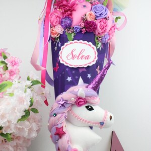 May include: A purple unicorn-themed gift cone decorated with pink, purple, and white flowers, ribbons, and a unicorn head. The cone has the name "Solea" on it. The unicorn has a sparkly horn and a braided mane.