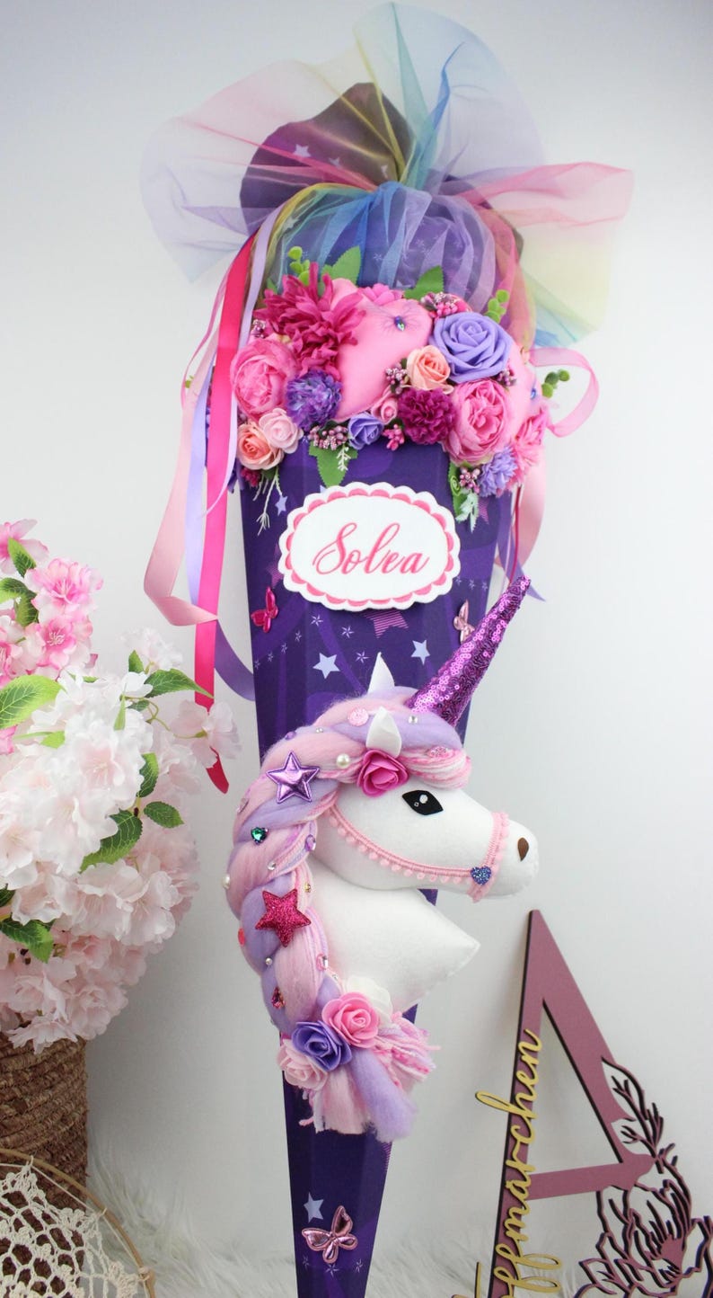 May include: A purple unicorn-themed gift cone decorated with pink, purple, and white flowers, ribbons, and a unicorn head. The cone has the name "Solea" on it. The unicorn has a sparkly horn and a braided mane.