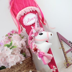 Könnte beinhalten: Eine rosa Schultüte mit Einhorn-Motiv, einem weißen Einhornkopf, einem paillettenbesetzten Horn und einem Namensschild mit der Aufschrift "Lilian". Die Tüte ist mit rosa Tüll, Bändern und Sternen verziert. Ein Korb mit rosa Blumen und ein Traumfänger befinden sich im Hintergrund.