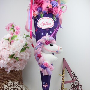 May include: A purple cone-shaped gift bag decorated with a unicorn head, flowers, and ribbons. The bag has a white label with the name "Solea" on it. The unicorn has a pink horn and a mane of pink and purple hair.