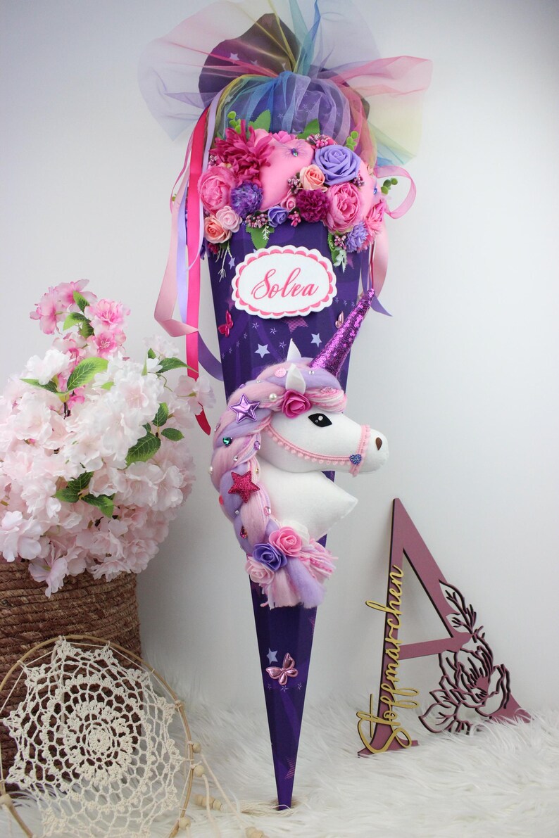 May include: A purple cone-shaped gift bag decorated with a unicorn head, flowers, and ribbons. The bag has a white label with the name "Solea" on it. The unicorn has a pink horn and a mane of pink and purple hair.