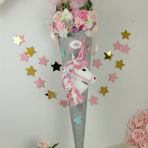 Könnte beinhalten: Ein grauer und rosa Einhorn-Schultüte mit einer rosa Schleife, Blumen und dem Namen "Ida" auf der Seite. Die Schultüte ist mit Sternen und Schmetterlingen verziert.