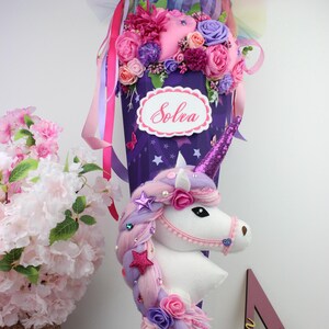 May include: A purple cone-shaped school bag decorated with a unicorn head, flowers, and ribbons. The bag has a white oval label with the name "Solea" in cursive. The unicorn has a pink horn and a mane of pink and purple hair.