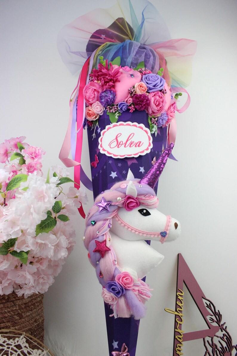 May include: A purple cone-shaped school bag decorated with a unicorn head, flowers, and ribbons. The bag has a white oval label with the name "Solea" in cursive. The unicorn has a pink horn and a mane of pink and purple hair.