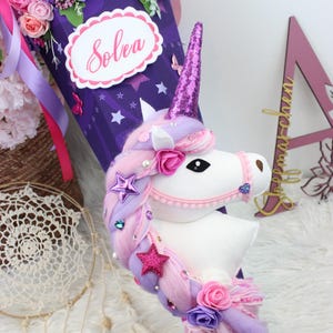 May include: A purple and pink unicorn-themed school cone decorated with flowers, stars, and butterflies. The cone features a white unicorn head with a pink horn and a label that says "Solea". The top is filled with pink and purple flowers.