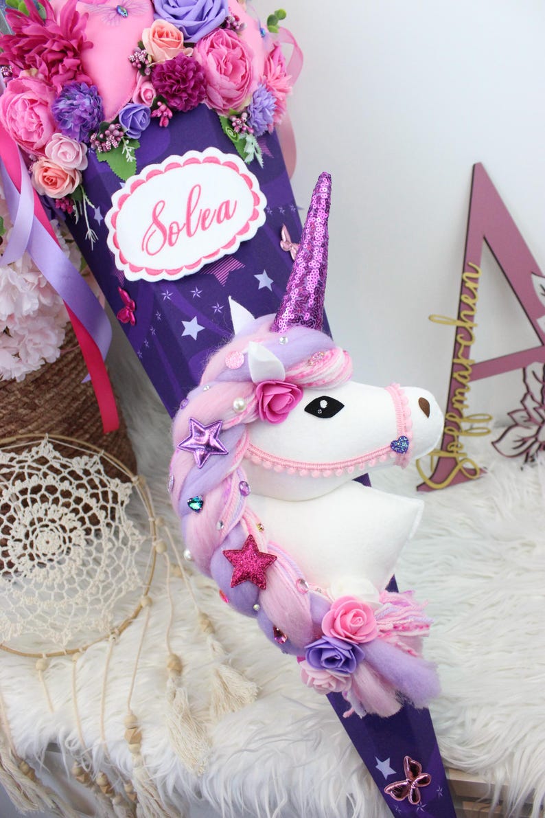 May include: A purple and pink unicorn-themed school cone decorated with flowers, stars, and butterflies. The cone features a white unicorn head with a pink horn and a label that says "Solea". The top is filled with pink and purple flowers.