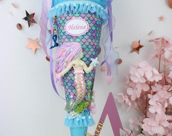 May include: A blue and purple mermaid-themed school cone with a pink and white mermaid doll, a blue tulle bow, and a name tag that says "Helene". The cone is standing on a pink wooden letter A with the word "Schultüten" written on it.