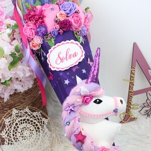 May include: A purple unicorn-themed school cone decorated with pink, purple, and white flowers, ribbons, and a plush unicorn head. The cone has the name "Solea" on it. The top is adorned with a rainbow tulle and more flowers.