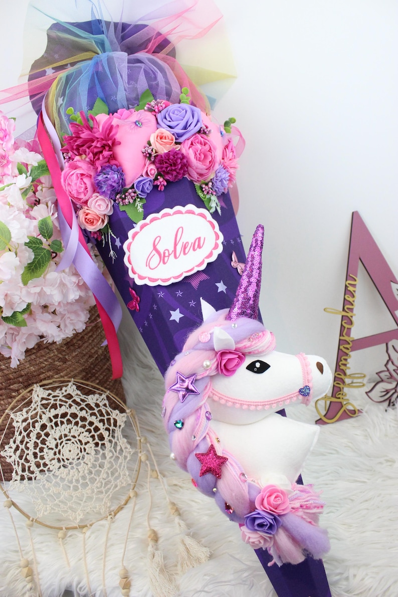 May include: A purple unicorn-themed school cone decorated with pink, purple, and white flowers, ribbons, and a plush unicorn head. The cone has the name "Solea" on it. The top is adorned with a rainbow tulle and more flowers.
