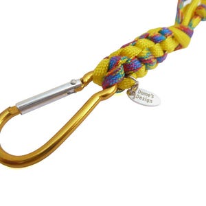 Op de afbeelding: Een gele en veelkleurige gevlochten lanyard met een gouden karabijnhaak. Een klein, ovaal label met de tekst "Dome's Design" is bevestigd. De lanyard is gemaakt van gevlochten nylon.