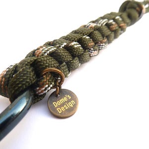 Peut inclure: Gros plan sur un porte-clés en paracorde tressée vert olive, marron et blanc. Une étiquette métallique de couleur bronze avec le texte "Dome's Design" est suspendue à un anneau en bronze. Un mousqueton en métal foncé est attaché.