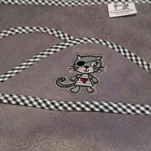 Könnte beinhalten: Ein grauer Filzstoff mit einem schwarz-weißen karierten Rand. In der Mitte des Stoffes befindet sich eine kleine gestickte Katze mit einem Piratenaugenklapp und einem roten Herzen auf ihrem Shirt. Der Text "AnuschkaDe handmade" befindet sich in der oberen rechten Ecke des Stoffes.