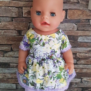 Könnte beinhalten: Eine Baby-Puppe, die ein weißes Kleid mit lila und gelbem Blumendruck und einem Lavendel-Spitzenbesatz trägt. Die Puppe trägt weiße Turnschuhe mit schwarzen Streifen.