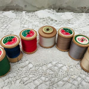 Könnte beinhalten: Eine Sammlung von Vintage Singer Nähgarnspulen in verschiedenen Farben, darunter Grün, Rot, Blau und Beige. Jede Spule hat einen Holzsockel und ein Papieretikett mit dem Singer-Logo und Text. Die Spulen sind auf einer weißen Spitzentischdecke angeordnet.