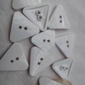 May include: A collection of white, triangular buttons with two holes each, scattered on a textured surface. Some buttons have a different back design with two holes. The buttons are made of a glossy material.