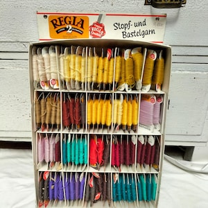 Może przedstawiać: Zabytkowe pudełko wystawowe wypełnione szpulkami przędzy marki Regia w różnych kolorach. Kolory przędzy obejmują odcienie żółtego, brązowego, czerwonego, turkusowego, fioletowego i różowego. Na pudełku widnieje napis "Regia" i "Stopf- und Bastelgarn".