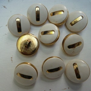 Peut inclure: Dix boutons blancs avec une bande dorée au centre. Les boutons sont ronds et ont une surface lisse.