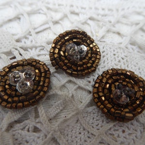 May include: Three round, spiral-shaped buttons. Each button is made of small, square, bronze-colored beads. The center of each button features a cluster of clear, faceted crystals, adding a touch of sparkle to the design. These buttons are ideal for embellishing clothing or craft projects.