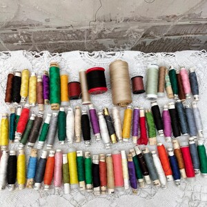 Könnte beinhalten: Eine Sammlung bunter Garnrollen, einige mit Etiketten, auf einer weißen Spitzentischdecke angeordnet. Die Rollen sind in verschiedenen Farbtönen von Rot, Blau, Grün, Gelb, Orange, Lila und Braun.