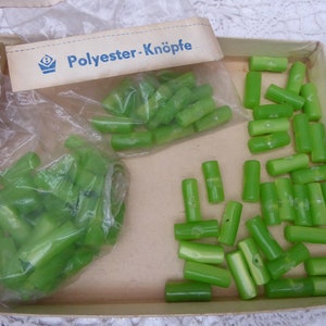 Peut inclure: Gros plan sur un paquet de boutons en plastique vert étiquetés "Polyester-Knöpfe". Les boutons sont dans deux sacs, l'un en plastique transparent et l'autre en carton brun.