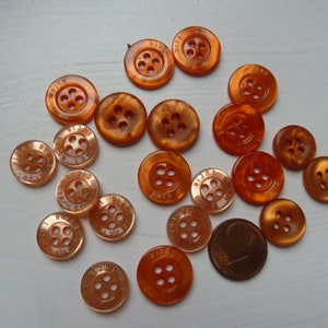 Op de afbeelding: Een verzameling van 18 oranje plastic knopen, elk met vier gaten. De knopen hebben een reliëfdesign en sommige hebben de tekst "BIG STAR" erop gedrukt.