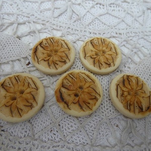 May include: Five wooden buttons with a floral design. The buttons are a light brown color with a darker brown floral pattern.