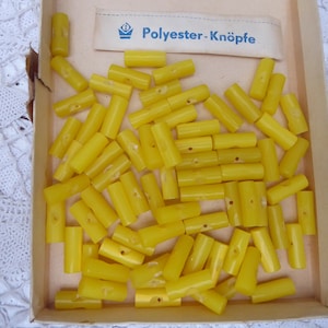 Pode incluir: Uma caixa de botões cilíndricos de poliéster amarelo brilhante, cada um com pequenos orifícios. A caixa tem um rótulo que diz "Polyester-Knöpfe". Os botões são de um amarelo vibrante, ideais para costura.