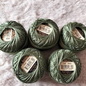 Könnte beinhalten: Fünf Knäuel grünes Anchor Pearl Cotton Garn, Col. 860, liegen auf einer weichen, weißen Oberfläche. Jedes Knäuel hat ein Etikett mit Produktinformationen und einem Barcode. Das Garn ist eng gewickelt und zeigt seine Textur und Farbe.
