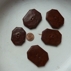 May include: Five brown, octagonal leather buttons with two holes each, arranged on a white surface. A copper penny is placed near one of the buttons for size comparison. The buttons have a stitched border.