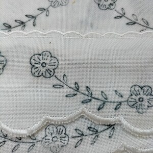 May include: Close-up of white fabric with a floral pattern. The fabric features a repeating design of stylized flowers and leaves in a dark gray color. The edges are scalloped with a white trim, creating a delicate and vintage aesthetic.