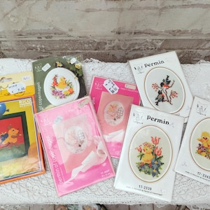 May include: A collection of cross-stitch kits featuring various designs, including birds, ducks, and floral arrangements. The kits are packaged in clear plastic and cardboard, with some displaying the "Permin" and "Rico Design" brands. The color palette includes yellow, pink, and green.